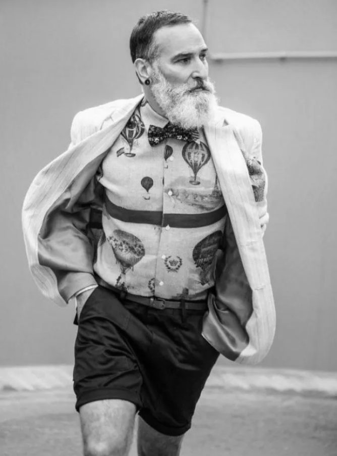 Hipsterzy — starszy mężczyzna z siwą brodą, w rozpiętej jasnej marynarce, koszuli w motywy balonów, muszce i krótkich spodenkach
